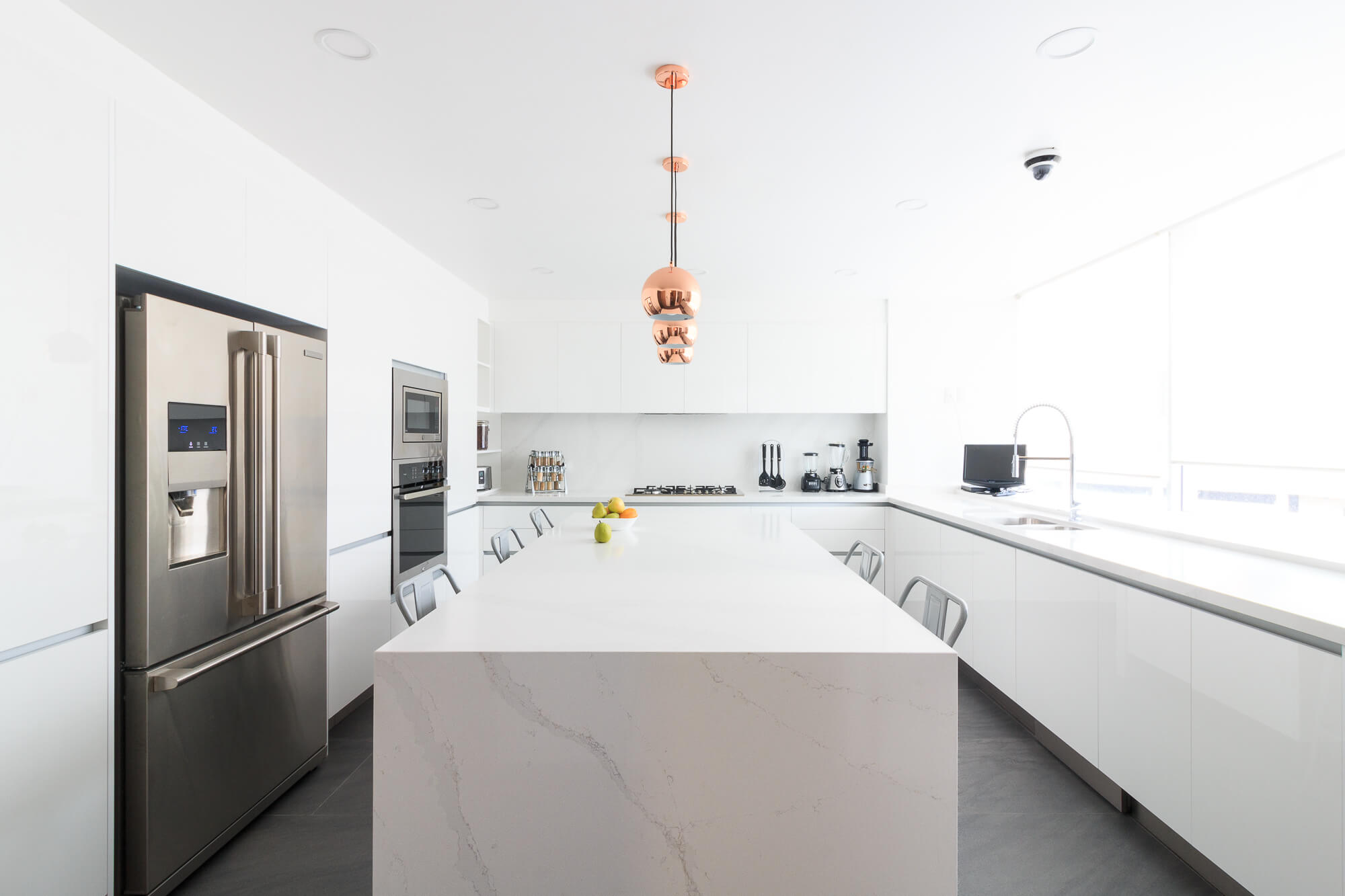 Cocina blanca con isla y cubierta tipo mármol y lámparas cobre.
