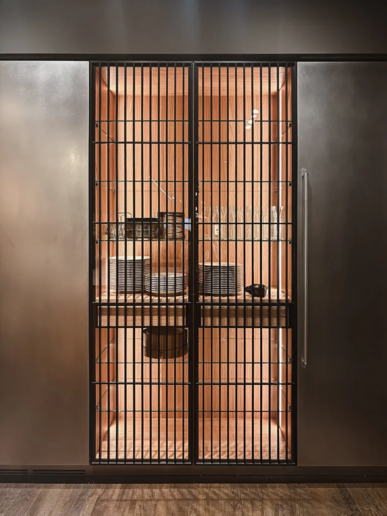 Vitrina alta de Boffi con frente de barrotes metálicos verticales en acabado oscuro e interior alistonado en madera clara, flanqueada por paneles laterales de acero mate. EuroCucina 2026.
