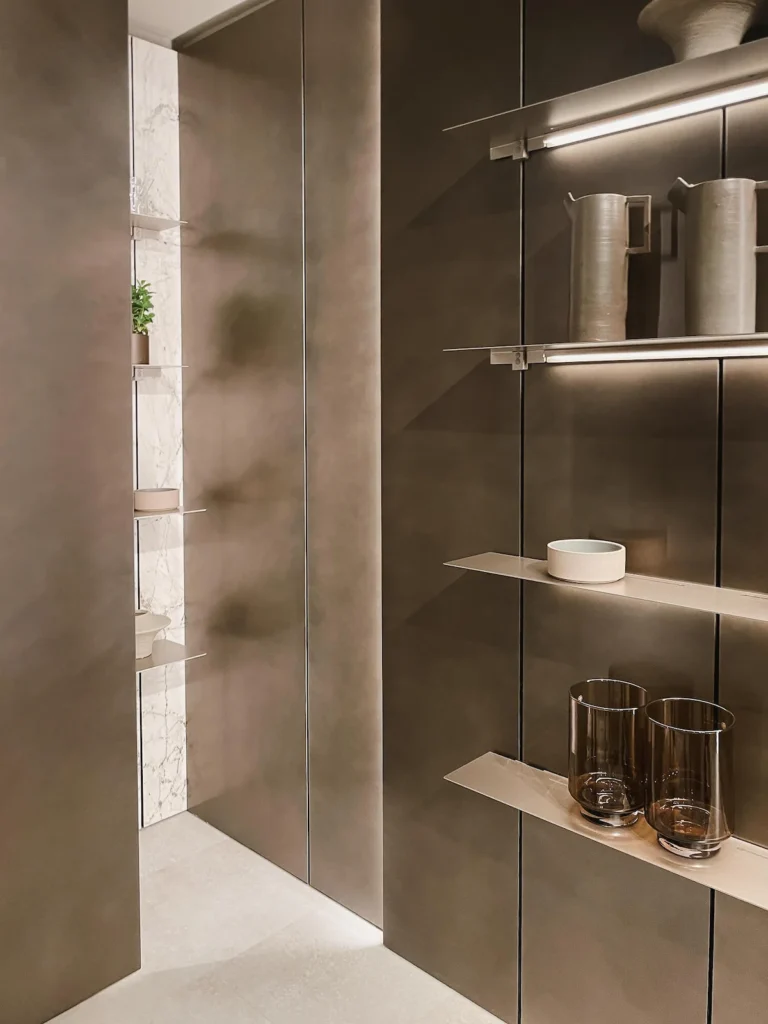 Paneles altos en acabado bronce claro satinado con repisas voladas iluminadas por luz rasante LED, junto a una hornacina revestida en mármol blanco veteado y bodegón en cerámica y cristal fumé, EuroCucina 2026.