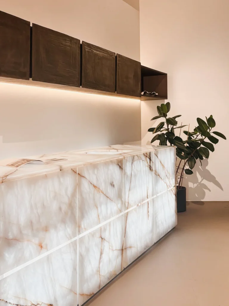 sla de la colección Terra de Minotti Cucine (Claudio Silvestrin) con envolvente en piedra Vitrum de Antolini retroiluminada — vetas cobrizas sobre fondo translúcido — bajo módulos altos colgados con puertas en placa de terracotta de canto irregular, EuroCucina 2026.