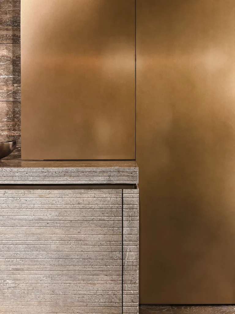 Detalle de cocina Modulnova en EuroCucina 2026: paneles altos de bronce satinado junto a frentes bajos en piedra acanalada y banda lateral de travertino veteado.
