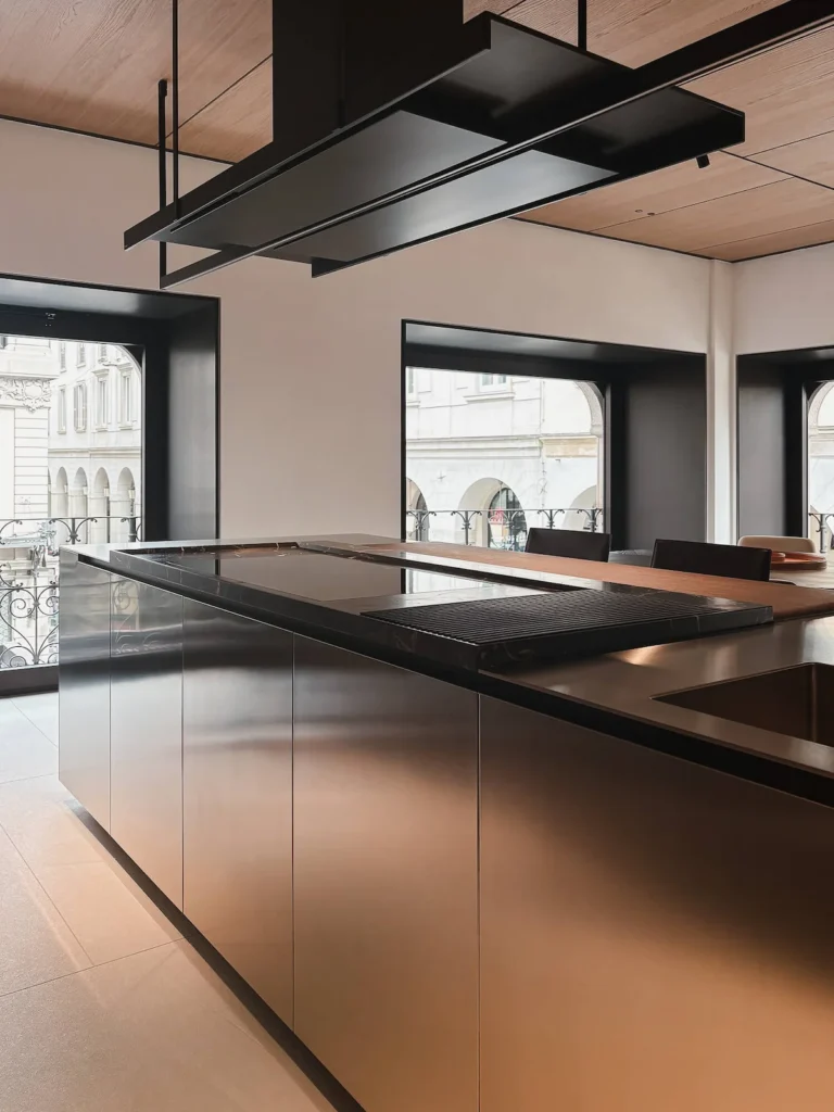 Isla de Poliform con cuerpo en acero inoxidable cepillado y cubierta combinada en mármol negro veteado — con placa de inducción y parrilla integradas — y madera oscura, bajo extractor suspendido en metal negro mate, EuroCucina 2026.
