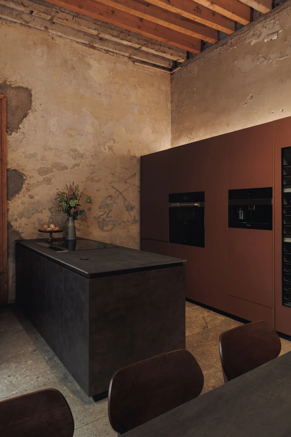 Casa Taittinger kitchen: aged metallic sheet island with 36-inch Miele induction cooktop facing the copper laminate appliance wall, over the original stone floor of Proyecto Público Prim.