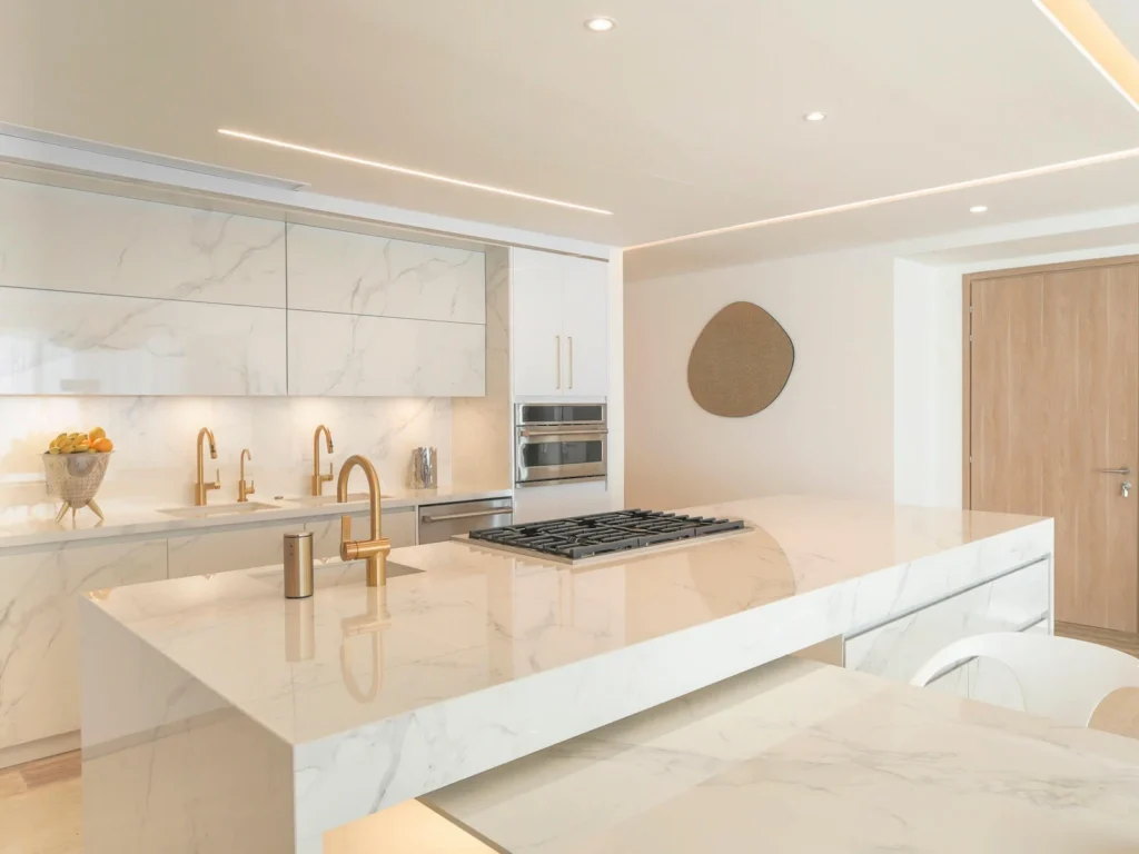 Vista frontal de cocina a medida con cubiertas y muros en Dekton Natura, horno empotrado y grifería dorada en Acapulco.